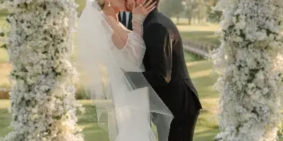 A bride and groom sharing a kiss under a white floral arch at an outdoor wedding venue.