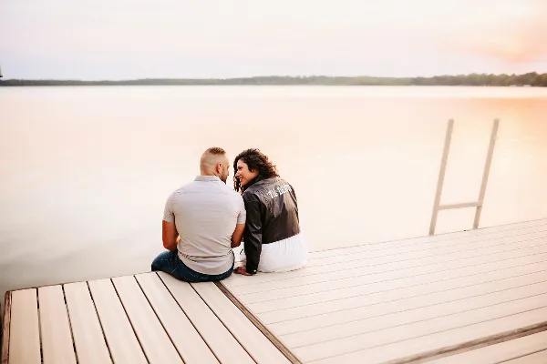 A couple sitting on a wooden dock by a lake at sunset.