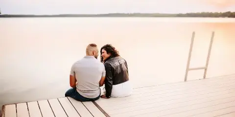 A couple sitting on a wooden dock by a lake at sunset.