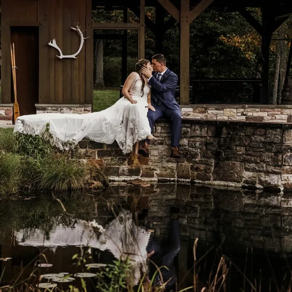 A bride and groom sitting on a stone ledge by a pond, sharing a kiss in a wedding portrait.