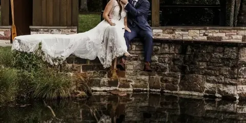 A bride and groom sitting on a stone ledge by a pond, sharing a kiss in a wedding portrait.