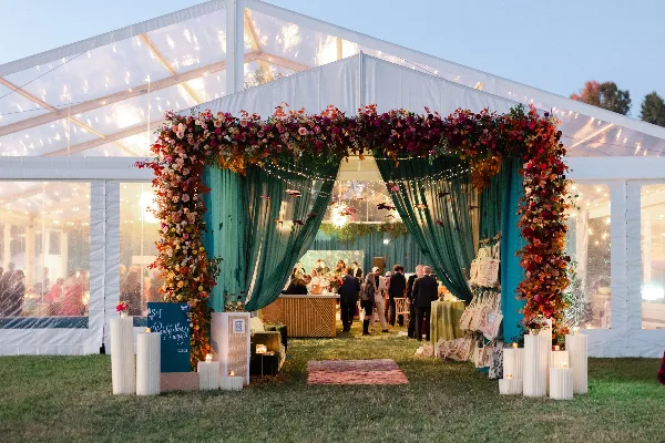 A wedding event entrance featuring a large floral archway and green drapery inside a clear tent.