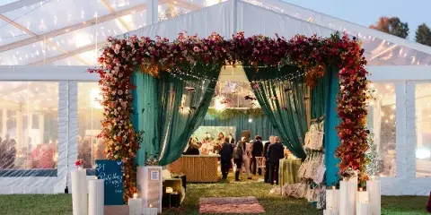 A wedding event entrance featuring a large floral archway and green drapery inside a clear tent.