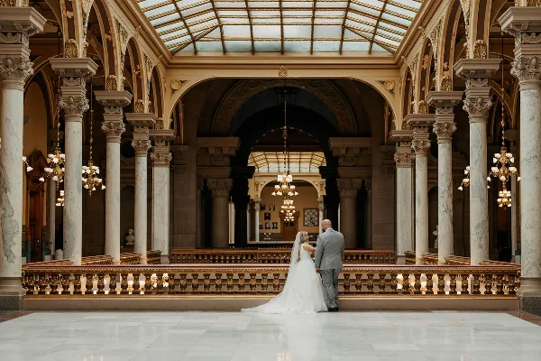 A bride and groom standing together in an ornate, columned hall with a glass ceiling.