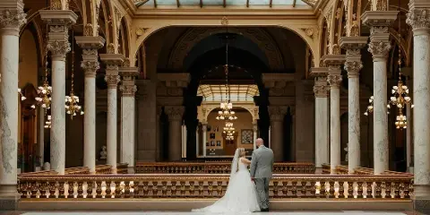A bride and groom standing together in an ornate, columned hall with a glass ceiling.