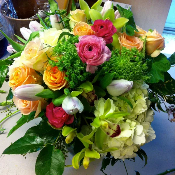 A lush floral arrangement containing peach roses, pink ranunculus, white tulips, green orchids, and white hydrangeas.