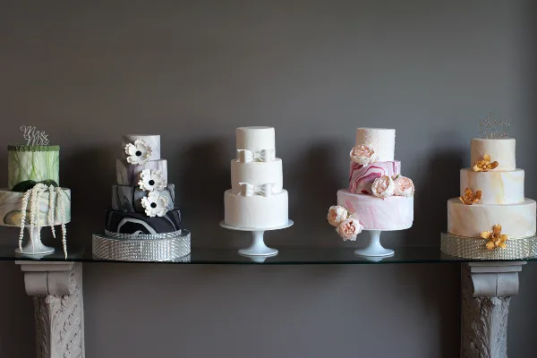 A row of five different custom wedding cakes displayed on a glass table against a dark background.