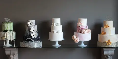 A row of five different custom wedding cakes displayed on a glass table against a dark background.