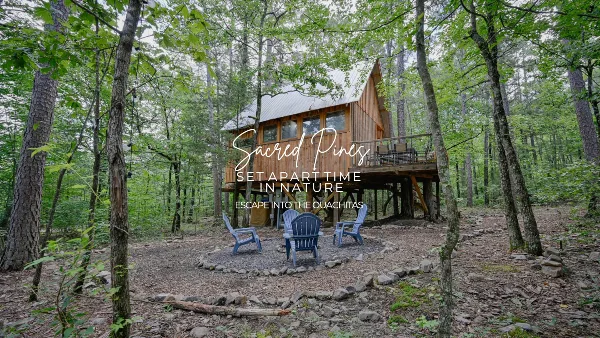 A wooden treehouse cabin in a forest with a fire pit and chairs.