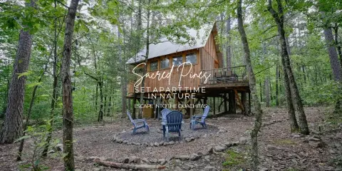 A wooden treehouse cabin in a forest with a fire pit and chairs.