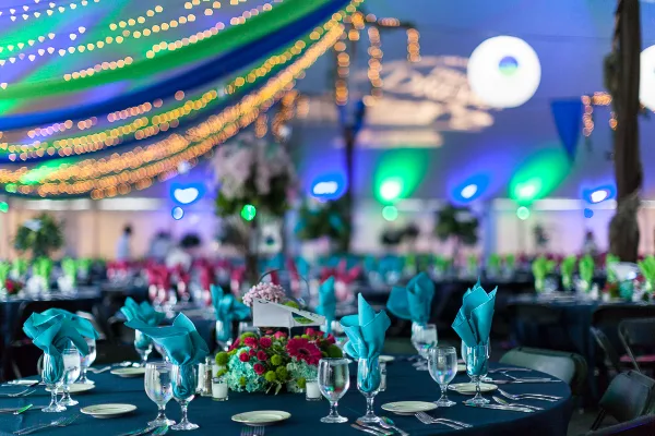 A decorated event table with teal napkins and glassware set up under a tent with string lights.