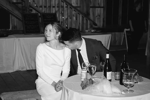 A bride and groom seated together at a reception table in a rustic wooden venue.