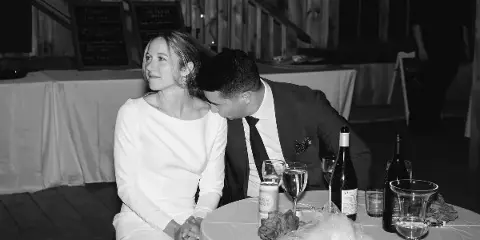 A bride and groom seated together at a reception table in a rustic wooden venue.