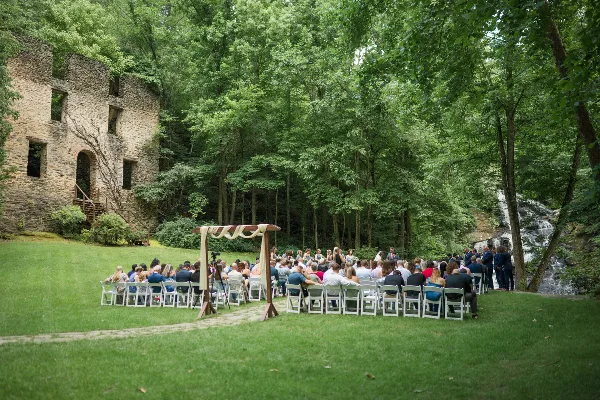 Outdoor wedding ceremony setup on a lawn with a stone gristmill ruin on the left and a waterfall in the background.