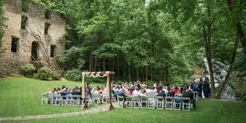 Outdoor wedding ceremony setup on a lawn with a stone gristmill ruin on the left and a waterfall in the background.