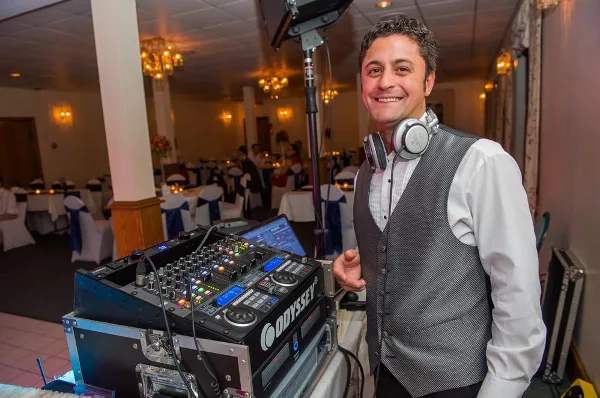 A smiling DJ in a vest with headphones around his neck stands beside a mixing console and laptop in a decorated wedding banquet hall.