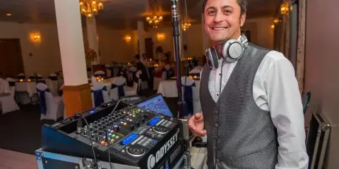 A smiling DJ in a vest with headphones around his neck stands beside a mixing console and laptop in a decorated wedding banquet hall.