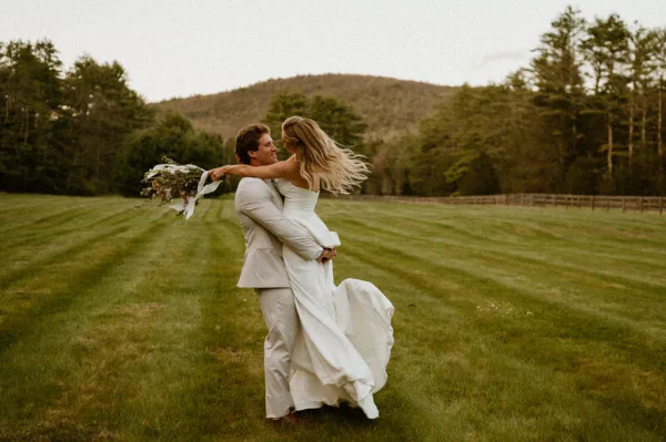 A bride and groom embracing and dancing in a grassy field with a forest background.