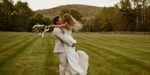 A bride and groom embracing and dancing in a grassy field with a forest background.