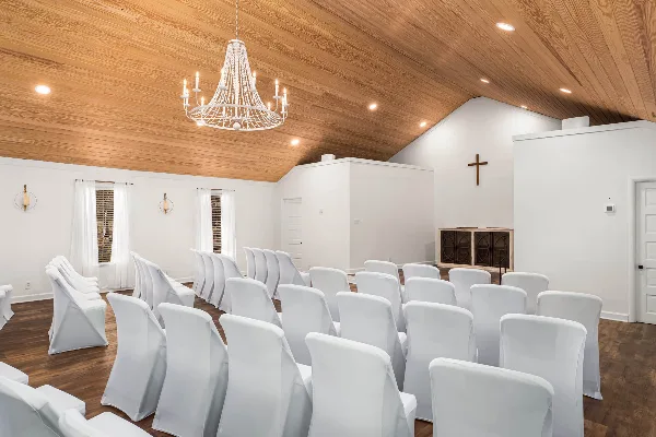 Interior view of a wedding chapel with white chairs, wooden ceiling, and a cross on the wall.