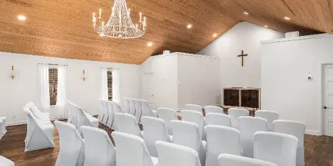 Interior view of a wedding chapel with white chairs, wooden ceiling, and a cross on the wall.
