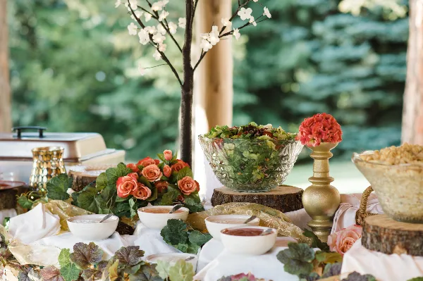 A catering buffet table set with a large glass bowl of salad, small white serving dishes, and floral arrangements on a white tablecloth.