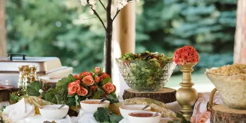 A catering buffet table set with a large glass bowl of salad, small white serving dishes, and floral arrangements on a white tablecloth.