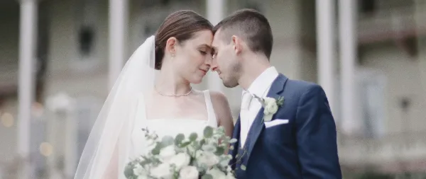 A bride and groom standing close together with foreheads touching, holding a white bouquet, in a cinematic wedding portrait.