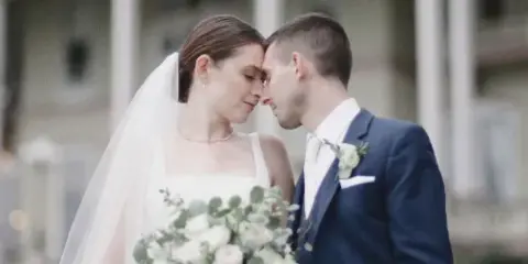A bride and groom standing close together with foreheads touching, holding a white bouquet, in a cinematic wedding portrait.