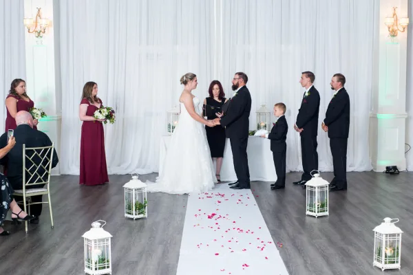 A bride and groom stand at the altar holding hands during a wedding ceremony, with an officiant standing between them and wedding party members on either side.