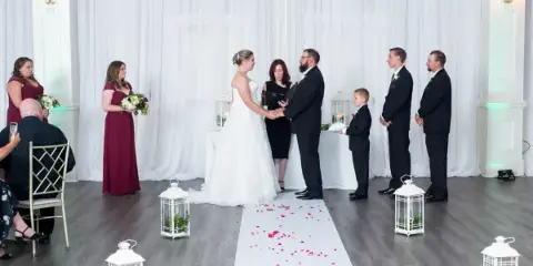 A bride and groom stand at the altar holding hands during a wedding ceremony, with an officiant standing between them and wedding party members on either side.