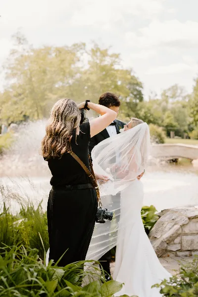 A photographer in black attire taking a picture of a bride and groom in a garden setting.