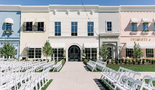 Exterior of a multi-story wedding venue with pastel storefronts, white ceremony chairs arranged on a lawn, and signage for Lyons Paperie and Desserts.