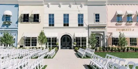 Exterior of a multi-story wedding venue with pastel storefronts, white ceremony chairs arranged on a lawn, and signage for Lyons Paperie and Desserts.