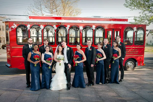 A wedding party posing in front of a red trolley decorated with white bows.