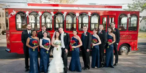 A wedding party posing in front of a red trolley decorated with white bows.