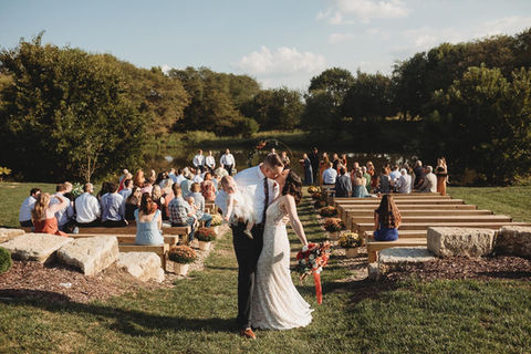 A bride and groom standing at an outdoor wedding ceremony on a grassy lawn with seated guests and wooden steps in the background.