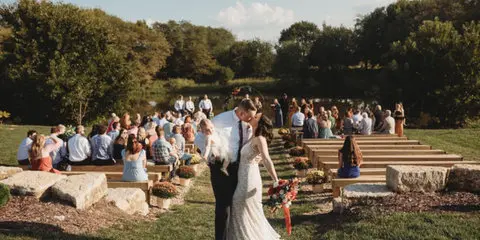 A bride and groom standing at an outdoor wedding ceremony on a grassy lawn with seated guests and wooden steps in the background.