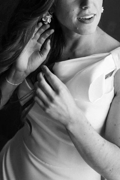 Black and white portrait of a smiling bride in a white dress, touching her hair with a delicate flower earring.