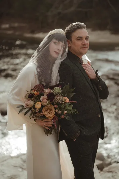 A bride in a white gown and veil holding a bouquet stands back-to-back with a groom in a dark suit near a body of water.