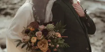 A bride in a white gown and veil holding a bouquet stands back-to-back with a groom in a dark suit near a body of water.