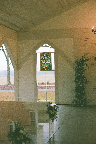 Interior view of a white chapel with a stained glass window, pews, and floral arrangements.