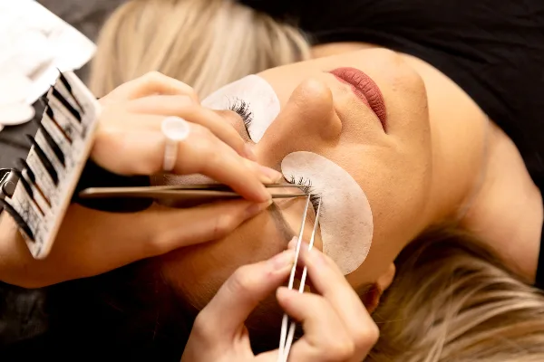 A close-up view of a beauty professional applying lash extensions to a client's eye using tweezers and a lash tray.