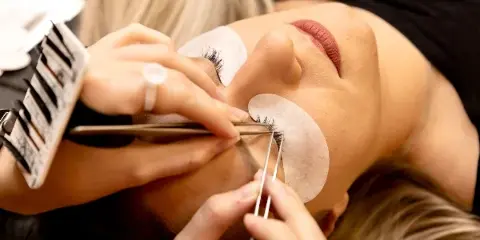 A close-up view of a beauty professional applying lash extensions to a client's eye using tweezers and a lash tray.