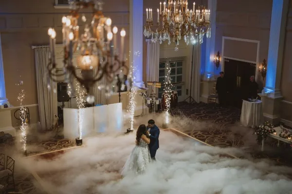 A bride and groom dancing on a fog-covered dance floor at a wedding reception with DJ equipment and chandeliers visible.