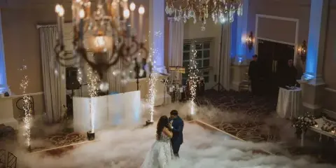 A bride and groom dancing on a fog-covered dance floor at a wedding reception with DJ equipment and chandeliers visible.