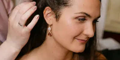 A bride in a white slip dress smiling softly while a hair stylist styles her hair.