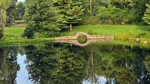 A stone arch bridge spanning a calm pond with a reflection of trees and greenery.