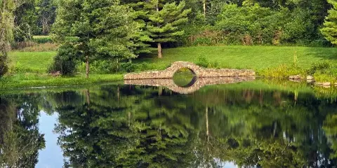A stone arch bridge spanning a calm pond with a reflection of trees and greenery.
