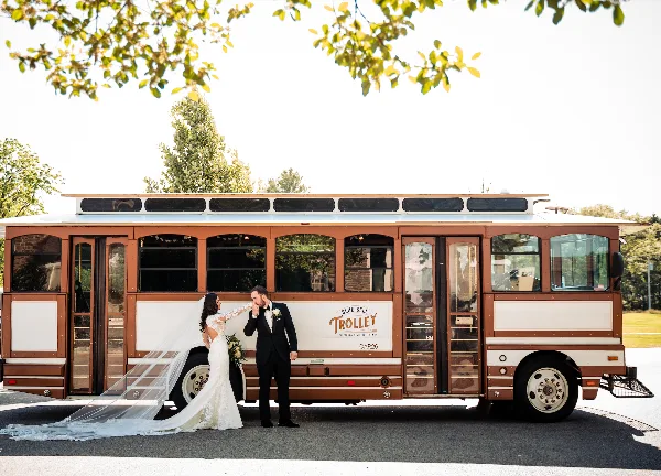 A bride and groom standing in front of a brown and white trolley bus with the Blue Bell Trolley logo.